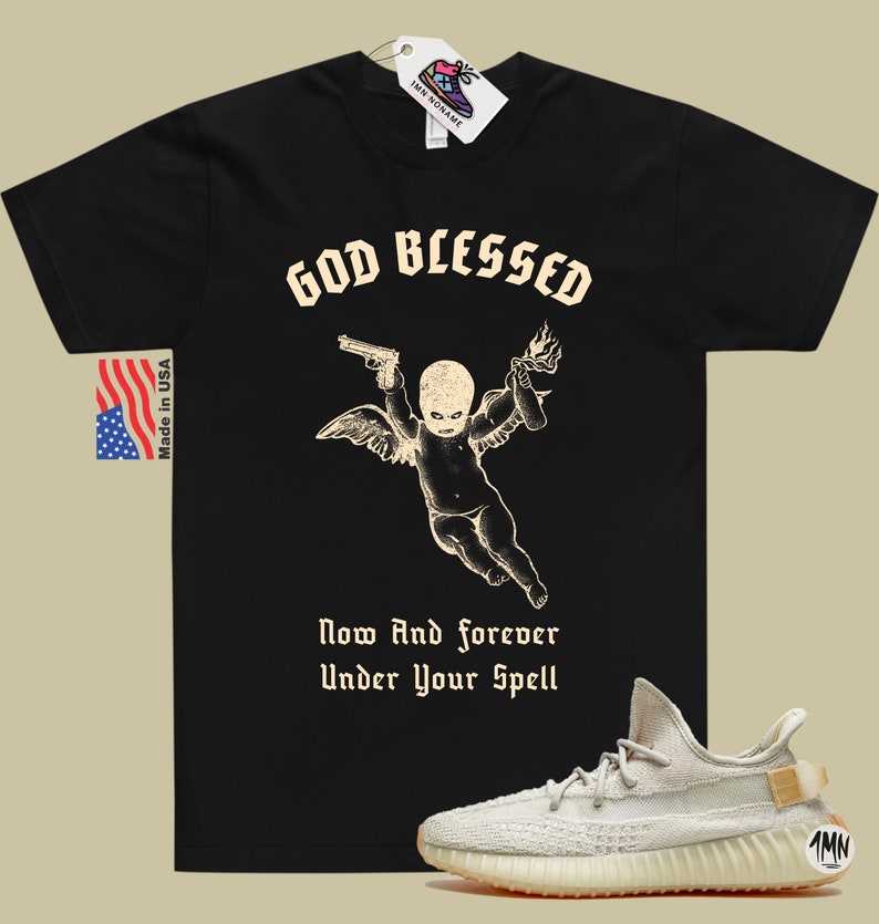 May include: Black t-shirt with a graphic of a cherub holding a gun and a Molotov cocktail. The text "God Blessed" is above the cherub and "Now And forever Under Your Spell" is below. A pair of light beige sneakers with a brown sole are in the bottom right corner of the image.