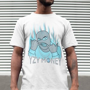 Puede incluir: Camiseta blanca con un gr&aacute;fico azul y gris de una persona con una m&aacute;scara de esqu&iacute; con llamas detr&aacute;s. El texto "YZY MONEY" est&aacute; impreso debajo del gr&aacute;fico.