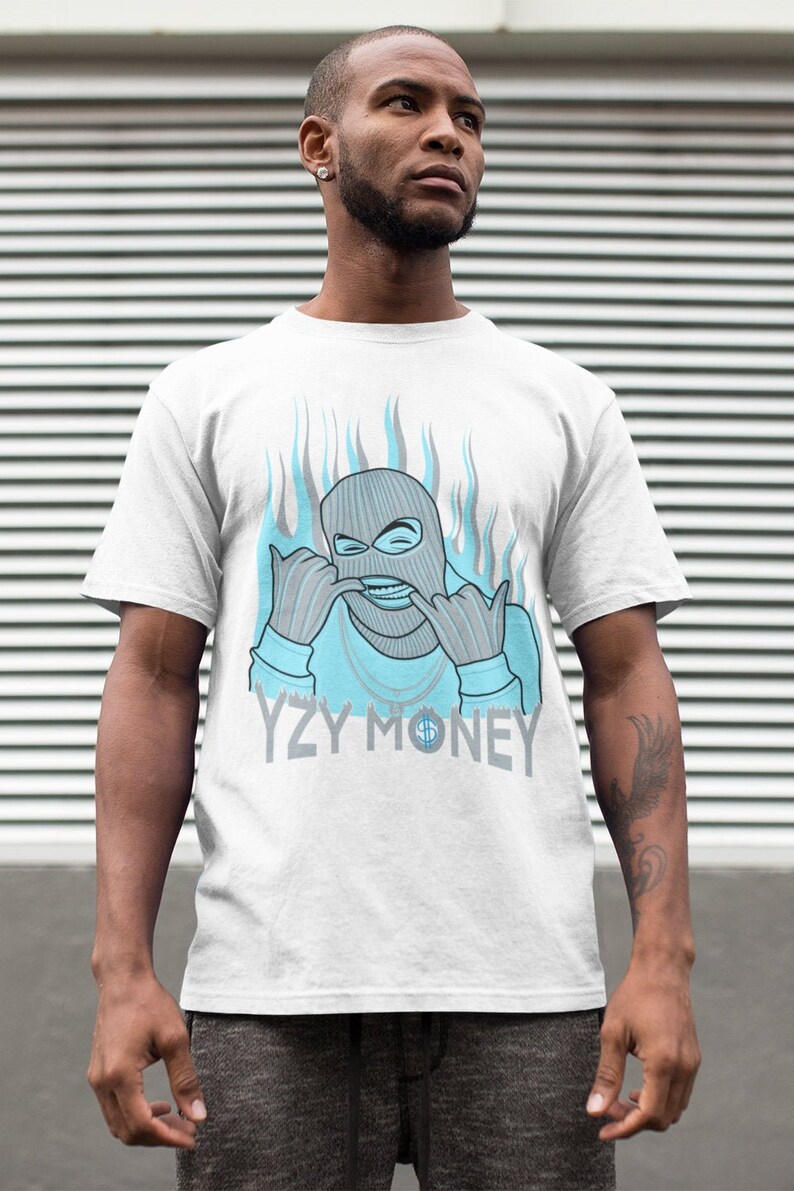 Puede incluir: Camiseta blanca con un gr&aacute;fico azul y gris de una persona con una m&aacute;scara de esqu&iacute; con llamas detr&aacute;s. El texto "YZY MONEY" est&aacute; impreso debajo del gr&aacute;fico.