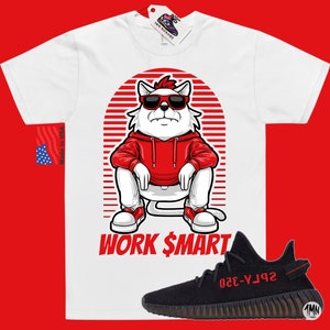 Puede incluir: Camiseta blanca con un gato de dibujos animados con gafas de sol y una sudadera roja. El gato est&aacute; sentado sobre un fondo a rayas rojo y blanco. El texto "WORK $MART" est&aacute; impreso debajo del gato. Un par de zapatillas Yeezy negras y rojas se muestran debajo de la camiseta.