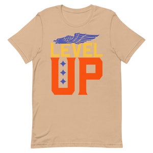 K&ouml;nnte beinhalten: Ein beiges T-Shirt mit kurzen &Auml;rmeln mit dem Text "LEVEL UP" in Orange und Gelb. Ein blauer Adler mit ausgebreiteten Fl&uuml;geln befindet sich &uuml;ber dem Text.