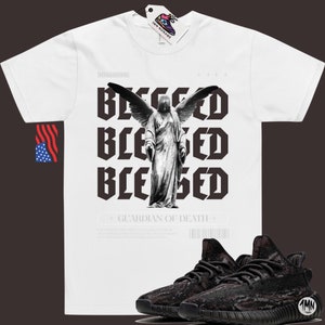 May include: White t-shirt with a black and white graphic of an angel with wings and the text "Blessed" repeated three times. The text "Guardian of Death" is below the graphic. A pair of black and brown sneakers are in the bottom right corner of the image.