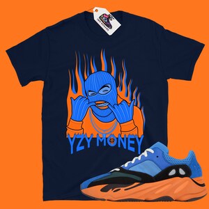 May include: A navy blue t-shirt with an orange and blue illustration of a person wearing a ski mask and a gold chain. The text "YZY MONEY" is printed below the illustration. A pair of blue and orange sneakers are shown in the bottom right corner of the image.