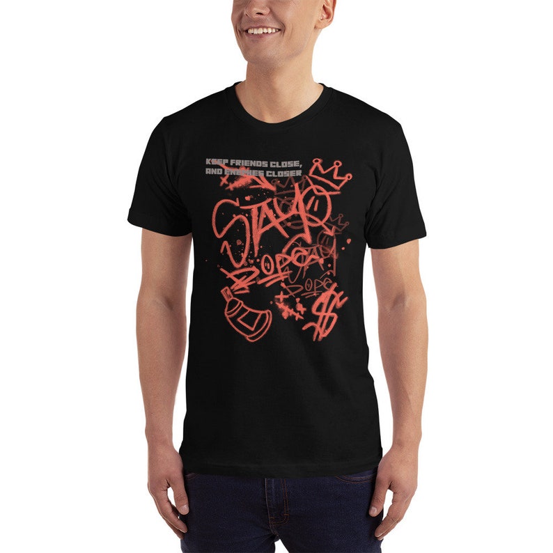 Puede incluir: Camiseta negra con un dise&ntilde;o gr&aacute;fico de pintura en aerosol roja. El dise&ntilde;o presenta las palabras "Stay" y "Dope" en un estilo graffiti. El texto "Keep friends close, and enemies closer" tambi&eacute;n est&aacute; incluido en el dise&ntilde;o.