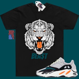 May include: Black t-shirt featuring a white lion graphic and the word "BEAST" in teal. The t-shirt is paired with white, grey, black and orange trainers with the text "Yeezy" on the side.