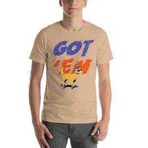 May include: A tan short-sleeved t-shirt with the text "GOT 'EM" in blue, orange, and white. A yellow cartoon dog wearing a blue hat and sunglasses is skateboarding.