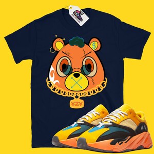 May include: Navy blue t-shirt featuring a cartoon bear design with gold chain and glasses, paired with yellow, orange, and blue sneakers. The bear graphic has the text "YZY" on a pendant. A tag with a shoe logo is attached.