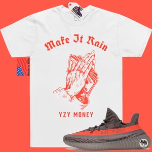 Puede incluir: Camiseta blanca con texto rojo que dice "Make It Rain" y un gr&aacute;fico de manos sosteniendo dinero. El texto "YZY MONEY" est&aacute; debajo del gr&aacute;fico. Un par de zapatillas Yeezy grises y naranjas est&aacute;n debajo de la camiseta.