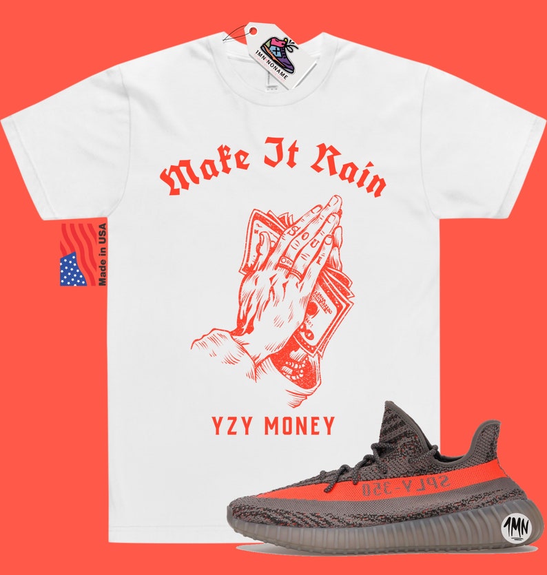 Puede incluir: Camiseta blanca con texto rojo que dice "Make It Rain" y un gr&aacute;fico de manos sosteniendo dinero. El texto "YZY MONEY" est&aacute; debajo del gr&aacute;fico. Un par de zapatillas Yeezy grises y naranjas est&aacute;n debajo de la camiseta.
