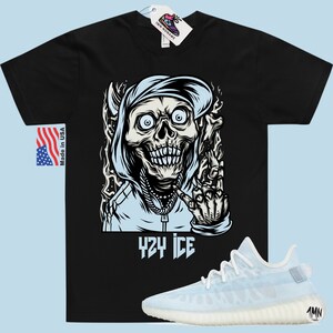 May include: Black t-shirt with a graphic of a skull wearing a blue hoodie and making a hand gesture. The text "424 ICE" is below the graphic. A pair of light blue and white sneakers are shown below the t-shirt.
