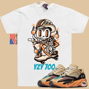 Puede incluir: Camiseta blanca con una ilustraci&oacute;n de dibujos animados de un esqueleto con un sombrero y montando un monopat&iacute;n. El texto "YZY 700" est&aacute; impreso debajo de la ilustraci&oacute;n. Un par de zapatillas Yeezy 700 de color marr&oacute;n claro y naranja se muestran en la esquina inferior derecha de la imagen.