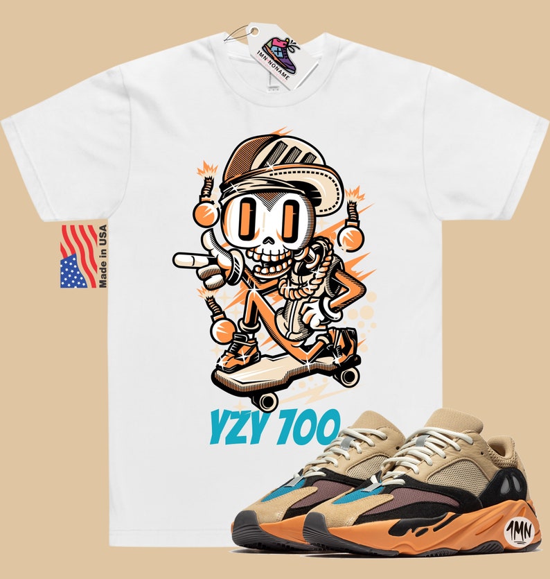 Puede incluir: Camiseta blanca con una ilustraci&oacute;n de dibujos animados de un esqueleto con un sombrero y montando un monopat&iacute;n. El texto "YZY 700" est&aacute; impreso debajo de la ilustraci&oacute;n. Un par de zapatillas Yeezy 700 de color marr&oacute;n claro y naranja se muestran en la esquina inferior derecha de la imagen.