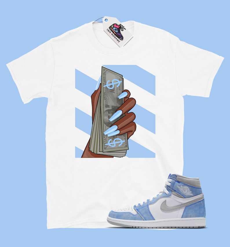 May include: White t-shirt featuring a graphic of a hand holding a stack of cash with blue dollar signs and long blue nails. The background is blue and white stripes. A pair of light blue and white trainers are also pictured.