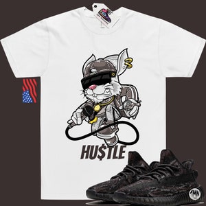 May include: White t-shirt with a cartoon rabbit wearing sunglasses, a gold chain, and a baseball cap. The rabbit is holding a microphone and a spray can. The text "HUSTLE" is printed below the rabbit. A pair of black and grey sneakers with a camouflage pattern are shown below the t-shirt.