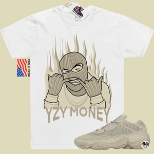 May include: A white t-shirt with a cartoon illustration of a person wearing a ski mask and a gold chain. The text "YZY MONEY" is printed below the illustration. A pair of tan Yeezy 500 sneakers are shown below the t-shirt.