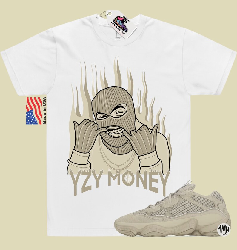 May include: A white t-shirt with a cartoon illustration of a person wearing a ski mask and a gold chain. The text "YZY MONEY" is printed below the illustration. A pair of tan Yeezy 500 sneakers are shown below the t-shirt.