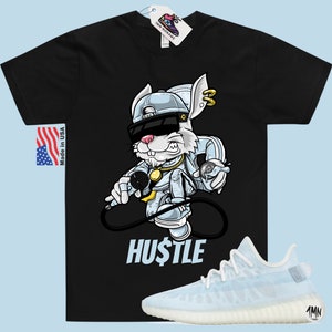May include: Black t-shirt featuring a cartoon rabbit wearing a backwards cap, sunglasses, and a gold chain. The rabbit is holding a microphone and a spray can. The text "HUSTLE" is printed below the rabbit. A pair of white and light blue trainers are shown below the t-shirt.