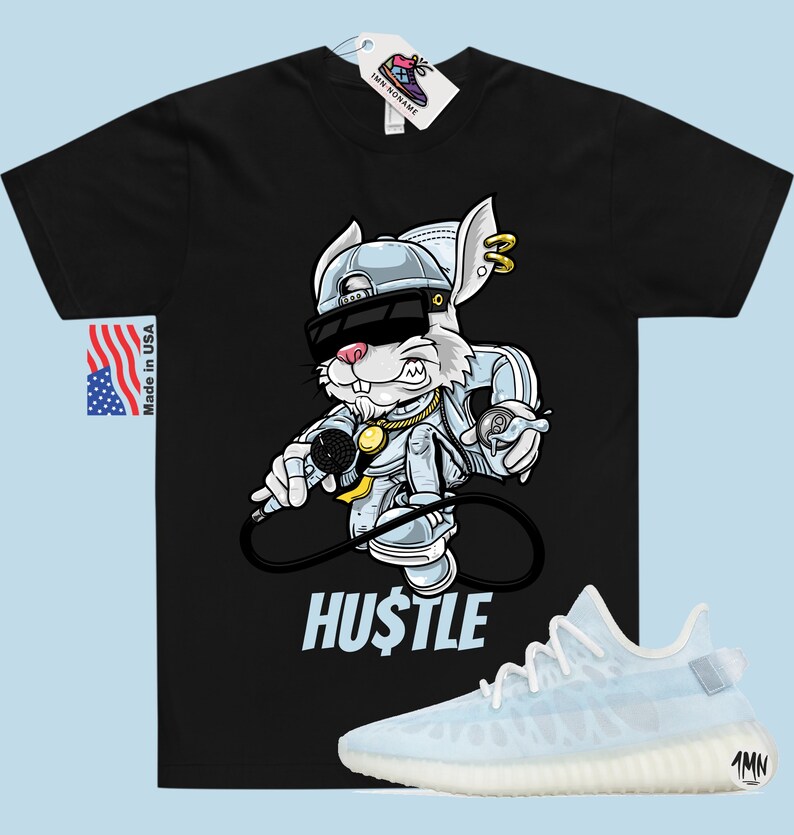 May include: Black t-shirt featuring a cartoon rabbit wearing a backwards cap, sunglasses, and a gold chain. The rabbit is holding a microphone and a spray can. The text "HUSTLE" is printed below the rabbit. A pair of white and light blue trainers are shown below the t-shirt.