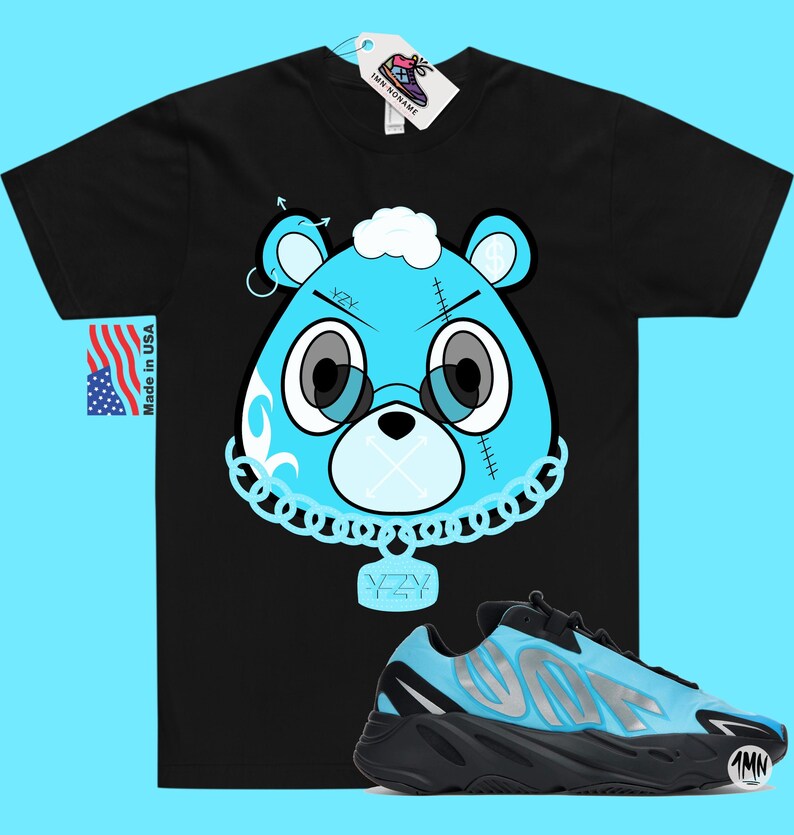 May include: Black t-shirt with a blue cartoon bear wearing a chain with a YZY pendant.  The bear has a stitched-up face and a cloud on its head.  A pair of blue and black YZY sneakers are below the t-shirt.