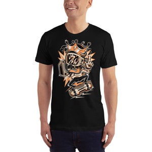 Może przedstawiać: Czarny T-shirt z kresk&oacute;wkową ilustracją postaci w koronie i okularach przeciwsłonecznych, trzymającej boombox i jeżdżącej na deskorolce. Postać jest pomarańczowa i biała, a tło jest czarne z pomarańczowymi i białymi akcentami.