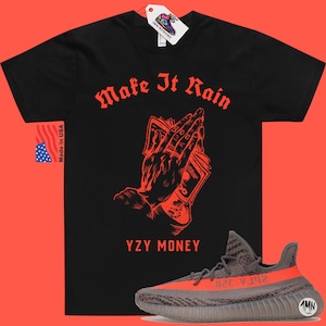 Puede incluir: Camiseta negra con texto rojo que dice "Make It Rain" y un gr&aacute;fico de una mano sosteniendo dinero. El texto "YZY MONEY" est&aacute; debajo del gr&aacute;fico. Un par de zapatillas grises y naranjas se encuentra en la esquina inferior derecha de la imagen.