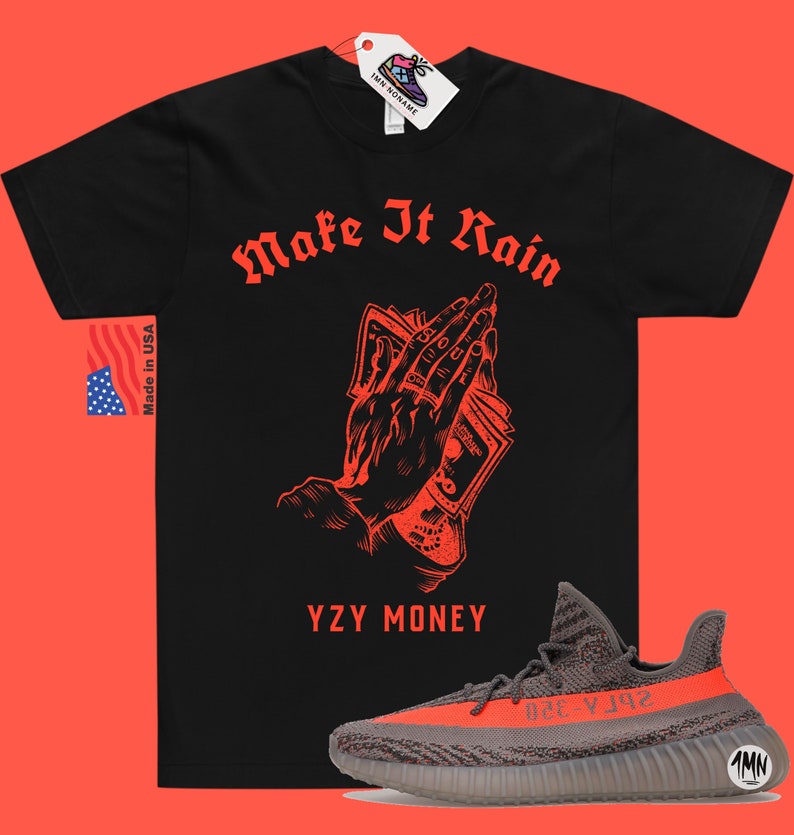 Puede incluir: Camiseta negra con texto rojo que dice "Make It Rain" y un gr&aacute;fico de una mano sosteniendo dinero. El texto "YZY MONEY" est&aacute; debajo del gr&aacute;fico. Un par de zapatillas grises y naranjas se encuentra en la esquina inferior derecha de la imagen.
