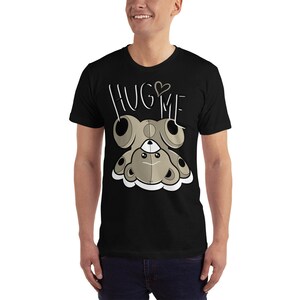 Puede incluir: Camiseta negra con un gr&aacute;fico de un oso de peluche marr&oacute;n con el texto "HUG ME" y un peque&ntilde;o coraz&oacute;n sobre la palabra "HUG".