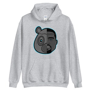 Peut inclure: Un sweat &agrave; capuche gris avec une illustration en noir et blanc d'une t&ecirc;te d'ours et du visage de Kanye West. La t&ecirc;te de l'ours est sur le c&ocirc;t&eacute; gauche de l'image et le visage de Kanye West est sur le c&ocirc;t&eacute; droit. Les deux visages sont s&eacute;par&eacute;s par une cl&ocirc;ture en fil barbel&eacute;. L'illustration est soulign&eacute;e en bleu.