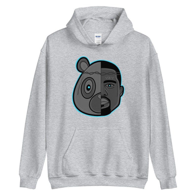 Peut inclure: Un sweat &agrave; capuche gris avec une illustration en noir et blanc d'une t&ecirc;te d'ours et du visage de Kanye West. La t&ecirc;te de l'ours est sur le c&ocirc;t&eacute; gauche de l'image et le visage de Kanye West est sur le c&ocirc;t&eacute; droit. Les deux visages sont s&eacute;par&eacute;s par une cl&ocirc;ture en fil barbel&eacute;. L'illustration est soulign&eacute;e en bleu.