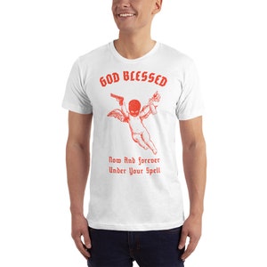 Puede incluir: Camiseta blanca con un gr&aacute;fico rojo de un querub&iacute;n con una m&aacute;scara de esqu&iacute;, sosteniendo una pistola y un c&oacute;ctel Molotov. El texto "GOD BLESSED" est&aacute; por encima del querub&iacute;n y "Now And forever Under Your Spell" est&aacute; por debajo.
