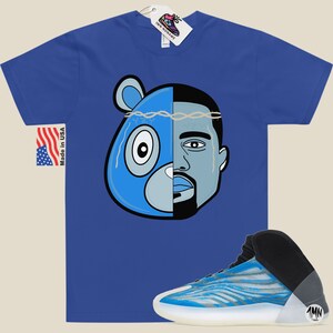 May include: A blue t-shirt with a graphic of a blue bear and Kanye West's face. The graphic has a barbed wire crown on top. The t-shirt is paired with a blue and white sneaker with a black upper and a white sole. The sneaker has a black tag with the letters "1MN" on it.