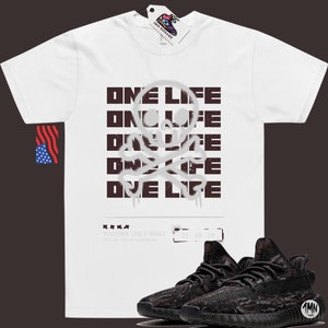 May include: White t-shirt with a black and white skull and crossbones graphic and the text "ONE LIFE" repeated five times. The text is in a graffiti style font. A pair of black and brown sneakers are also pictured.
