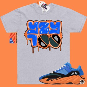 May include: A grey t-shirt with a blue and orange graphic that says "#YZY 700" with a pair of sunglasses. The t-shirt is paired with a blue, orange, and black sneaker.
