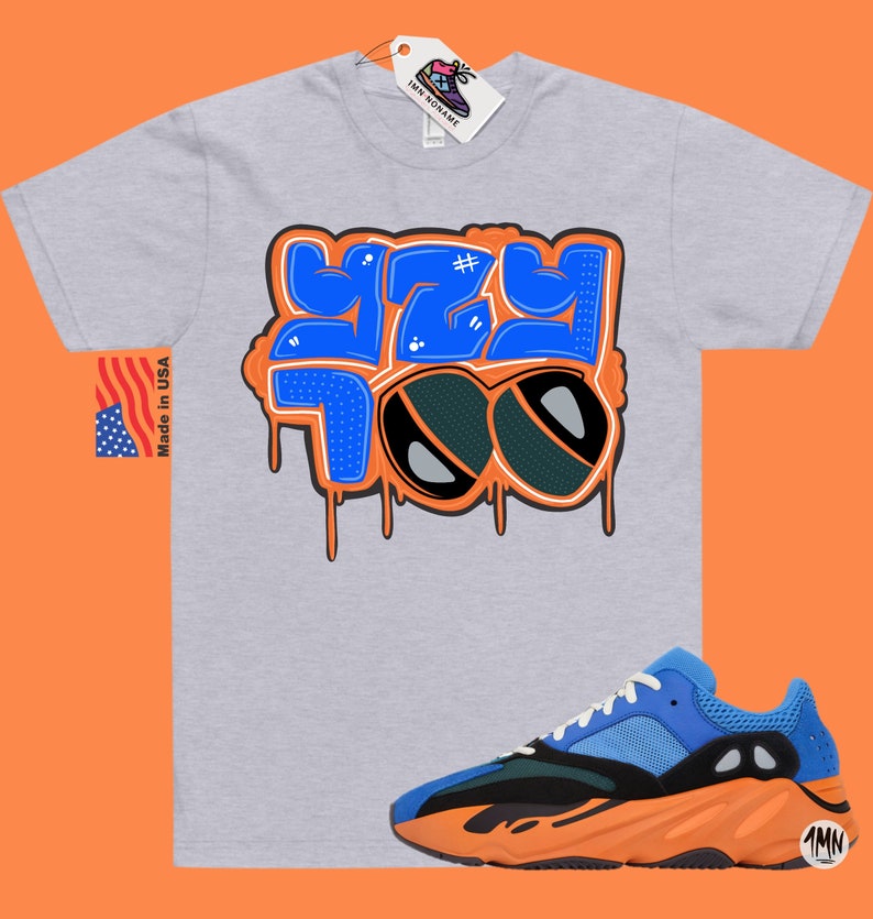 May include: A grey t-shirt with a blue and orange graphic that says "#YZY 700" with a pair of sunglasses. The t-shirt is paired with a blue, orange, and black sneaker.