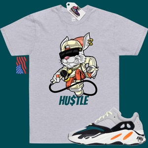 May include: A grey t-shirt featuring a cartoon rabbit wearing sunglasses, a hat, and a gold chain. The rabbit is holding a microphone and a spray can. The text "HUSTLE" is printed below the rabbit. A pair of white, green, and orange trainers are shown below the t-shirt.