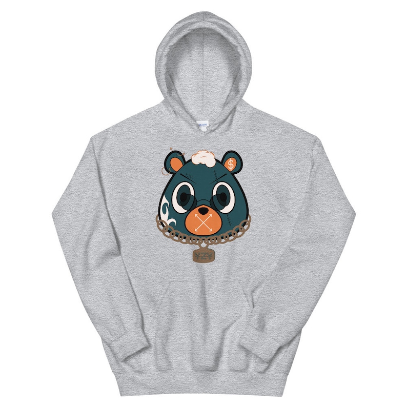 May include: A light gray hooded sweatshirt featuring a cartoon bear face. The bear has a teal face with large white eyes, an orange mouth, and a gold chain necklace. The bear's ears are orange with dollar signs. The tag on the necklace says "YZY".