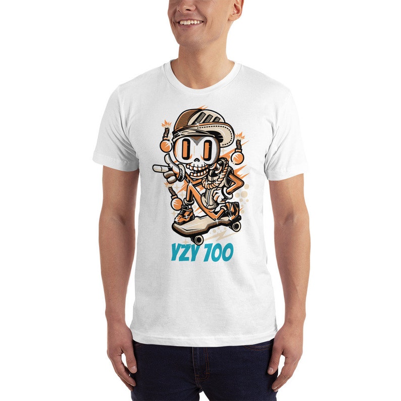 Puede incluir: Camiseta blanca con una ilustraci&oacute;n de dibujos animados de un esqueleto haciendo skateboard. El esqueleto lleva un sombrero, gafas de sol y un pa&ntilde;uelo. El texto "YZY 700" est&aacute; impreso debajo de la ilustraci&oacute;n.