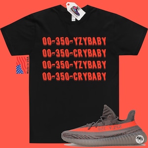 Peut inclure: T-shirt noir avec le texte "00-350-YZBABY" et "00-350-CRYBABY" r&eacute;p&eacute;t&eacute; en rouge. Une paire de baskets grises et orange avec le texte "350-YZY" sur le c&ocirc;t&eacute;.