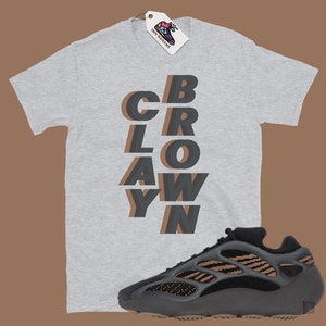 May include: A light gray t-shirt with the word "CBRLOAWYN" in a layered, shadow effect design. A black and brown athletic shoe is positioned below the shirt. A tag with a shoe graphic hangs from the shirt.