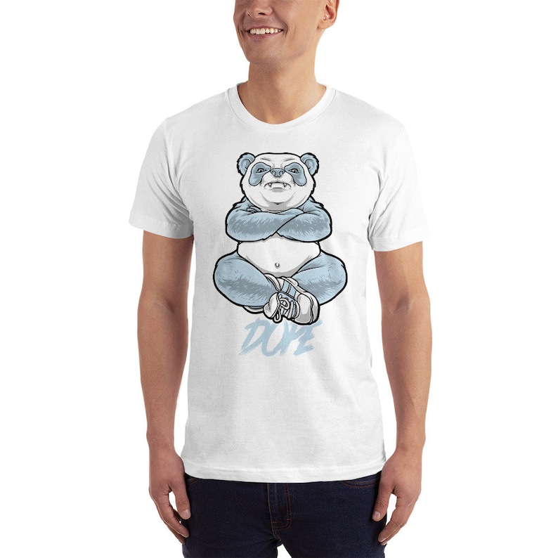 May include: White t-shirt featuring a blue panda bear sitting with its arms crossed and the word "DUPE" printed below.
