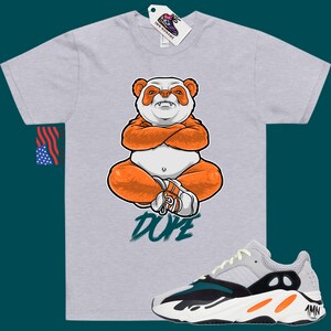 Pu&ograve; includere: Una maglietta grigia con un panda dei cartoni animati che indossa abiti arancioni e la parola "DOPE" stampata sul davanti. Un paio di scarpe da ginnastica bianche, nere, arancioni e turchesi sono mostrate sotto la maglietta.