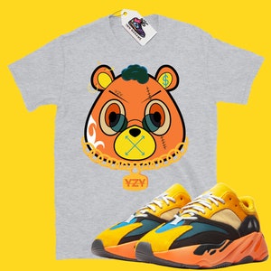 May include: A gray t-shirt featuring a cartoon bear design with glasses, a dollar sign, and the letters "YZY". Paired with yellow, orange, and blue sneakers. A tag with the text "IMN NONAME" is attached to the shirt.