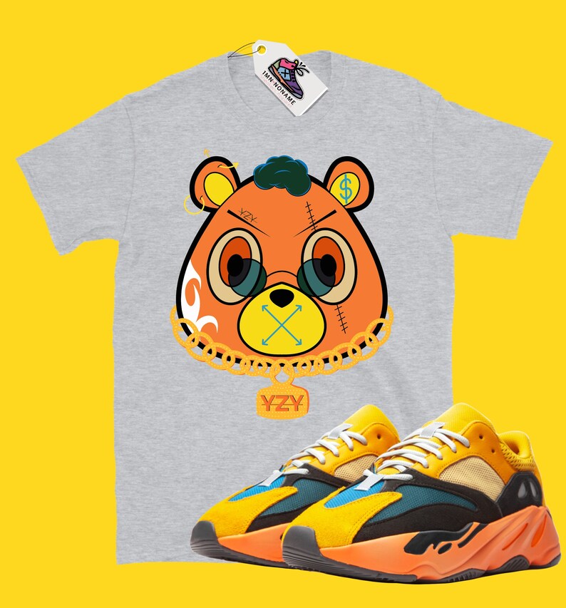 May include: A gray t-shirt featuring a cartoon bear design with glasses, a dollar sign, and the letters "YZY". Paired with yellow, orange, and blue sneakers. A tag with the text "IMN NONAME" is attached to the shirt.