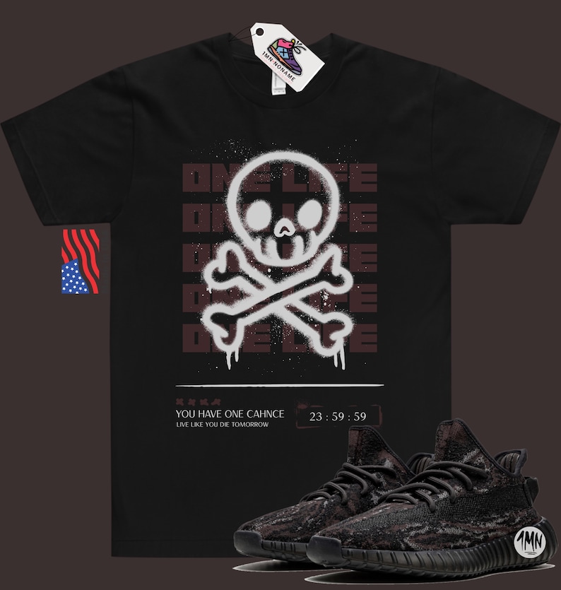 May include: Black t-shirt with a white skull and crossbones graphic. The graphic is a spray paint design with the word "ONE LIFE" repeated in a faded brown color. The text "YOU HAVE ONE CHANCE LIVE LIKE YOU DIE TOMORROW 23:59:59" is printed below the graphic. A pair of black and grey sneakers are shown below the t-shirt.