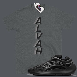 Puede incluir: Una camiseta gris con la palabra "ALVAH" impresa verticalmente en una fuente superpuesta, gris y negra. Una zapatilla negra y gris se muestra debajo de la camiseta.
