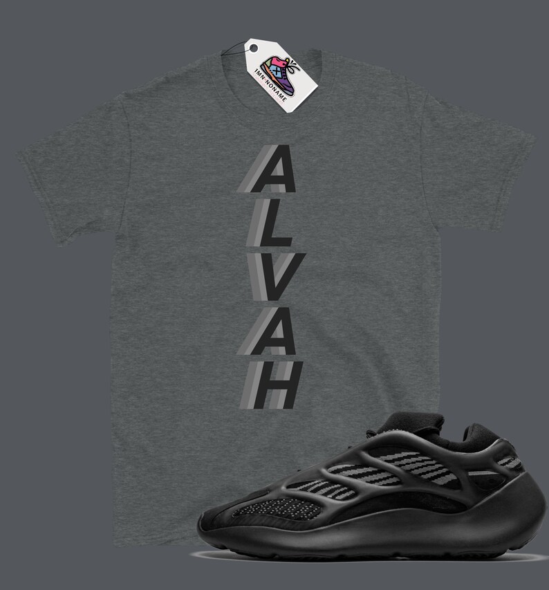 Puede incluir: Una camiseta gris con la palabra "ALVAH" impresa verticalmente en una fuente superpuesta, gris y negra. Una zapatilla negra y gris se muestra debajo de la camiseta.