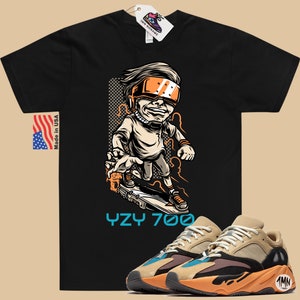 May include: Black t-shirt with a cartoon illustration of a person wearing a futuristic helmet and riding a skateboard. The text "YZY 700" is printed below the illustration. A pair of tan, orange, and blue sneakers are shown below the t-shirt.