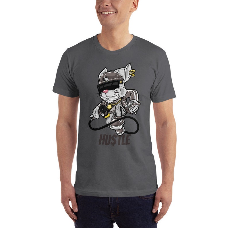 May include: A dark gray t-shirt with a cartoon rabbit wearing a backwards baseball cap, sunglasses, and a gold chain. The rabbit is holding a microphone and a spray can. The text "HUSTLE" is printed below the rabbit.