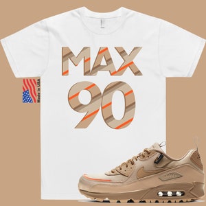 May include: A white t-shirt with the text "MAX 90" in brown and orange stripes. A pair of tan Nike Air Max 90 trainers with orange accents.