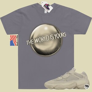 May include: A gray t-shirt with the text "The World Is Yours" printed on a gold sphere. A pair of beige sneakers with a mesh upper and a thick sole are shown below the t-shirt.